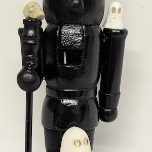 May include: A black wooden nutcracker in the form of a king, wearing a crown and holding a staff. It features a skull and two ghost figures. This is a Halloween or gothic-themed decorative item.