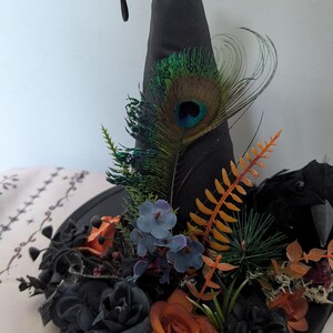 May include: Black witch's hat decorated with flowers, peacock feathers, and a raven. The hat has a silver bird skull accent and a black cord. The base is adorned with orange, blue, and black floral elements.
