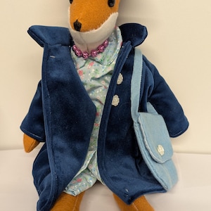 May include: A plush fox doll dressed in a blue hat, coat, and handbag. The fox has an orange face and legs, with a floral print shirt and a pink beaded necklace. The hat has a light blue band and a pink flower.
