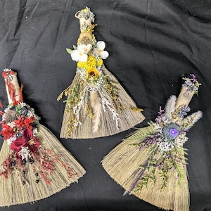 May include: Three decorative brooms with natural straw bristles and wooden handles. Each broom is decorated with dried flowers and foliage in shades of red, yellow, purple, and white. The brooms are arranged on a black surface.