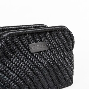 May include: Close-up of a black woven handbag with a textured, chevron-like pattern. The bag features a rectangular label with the brand name "CHIELLE". The top edge is smooth and rounded, and the bag appears to be made of a durable material.