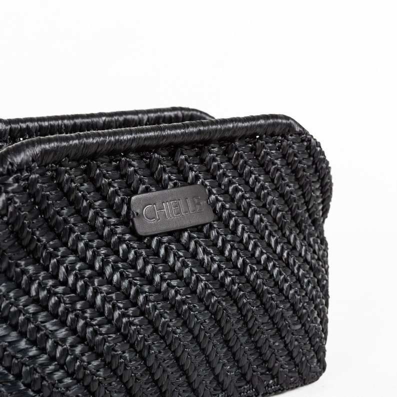May include: Close-up of a black woven handbag with a textured, chevron-like pattern. The bag features a rectangular label with the brand name "CHIELLE". The top edge is smooth and rounded, and the bag appears to be made of a durable material.
