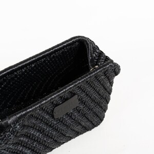May include: A black woven handbag with a rectangular shape. The bag features a textured surface and a small, rectangular leather tag with white text. The bag is set against a white background.