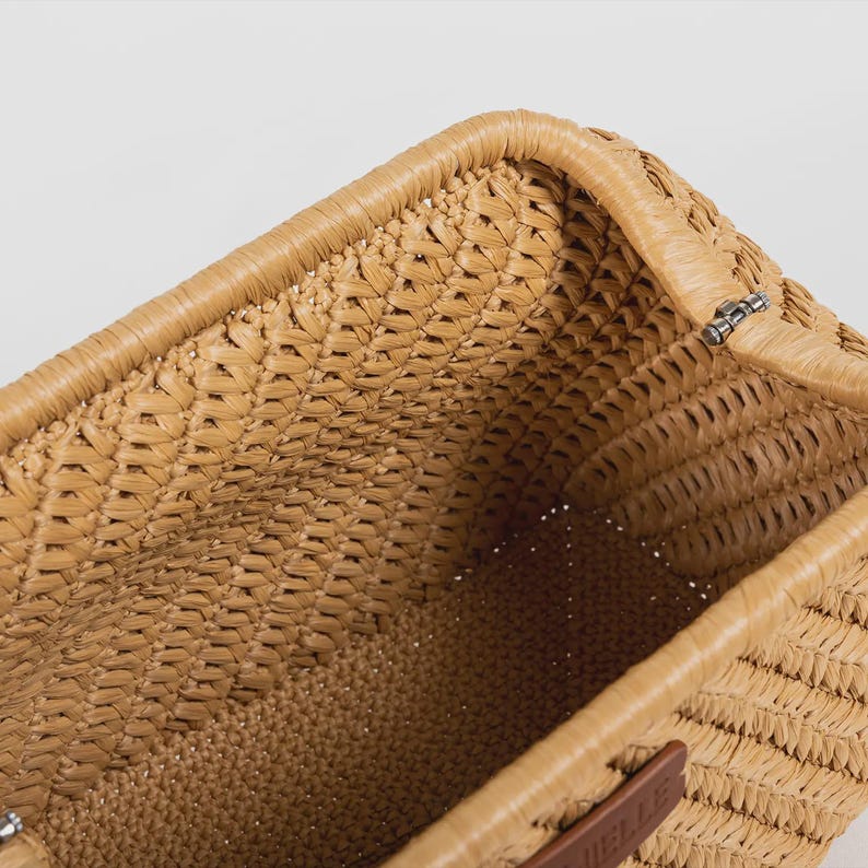May include: Close-up of a woven straw tote bag. The bag is a light tan color with a rectangular shape. The weave pattern is visible, and the edges are reinforced with a thicker straw material. A small leather tag is visible.