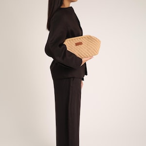 May include: A woven beige clutch with a rectangular shape and a brown leather label. The clutch is held against a person wearing a dark brown blazer and matching pants. The background is a plain white.