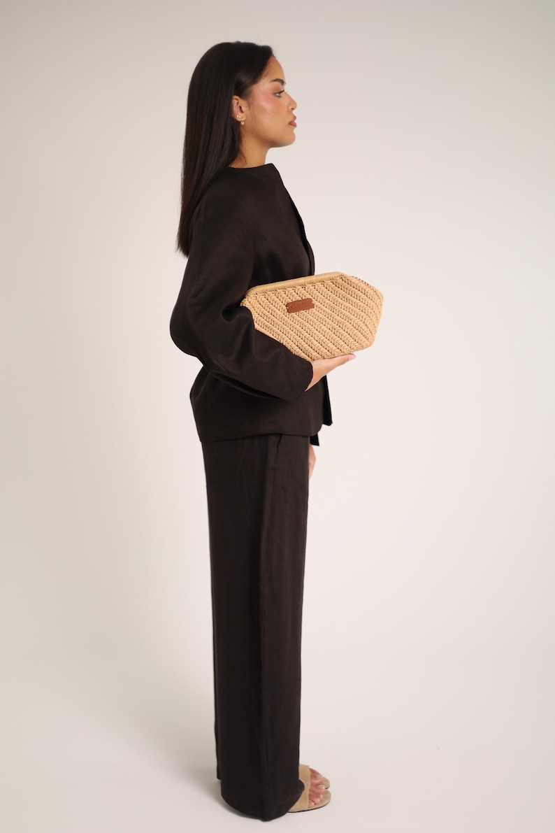 May include: A woven beige clutch with a rectangular shape and a brown leather label. The clutch is held against a person wearing a dark brown blazer and matching pants. The background is a plain white.