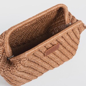 May include: A woven brown clutch bag with a rectangular shape and a textured, open interior. The bag features a woven pattern and a small brown leather tag. The bag is open, revealing the interior.