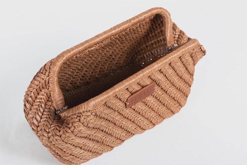 May include: A woven brown clutch bag with a rectangular shape and a textured, open interior. The bag features a woven pattern and a small brown leather tag. The bag is open, revealing the interior.