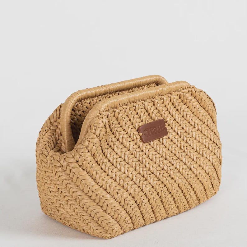 May include: A woven straw handbag in a light tan color. The bag has a rectangular shape with a curved top and two handles. A small brown leather tag with the word "CHELIE" is attached to the front.