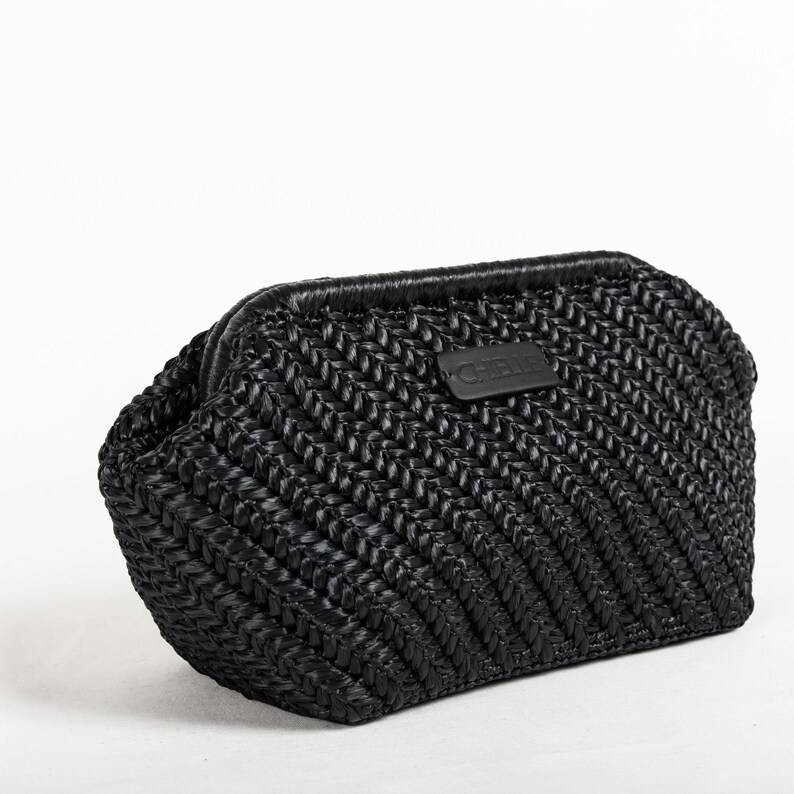 May include: Black woven clutch with a chevron pattern. The bag has a rectangular shape with rounded edges and a top closure. A small, rectangular leather patch with the brand name is centered on the front.