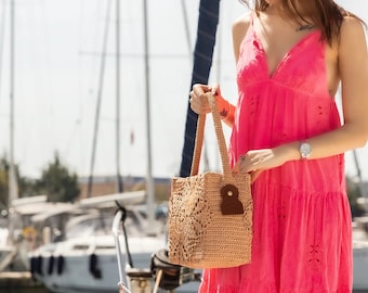 May include: A woman in a pink dress holds a crocheted tote bag with a brown leather accent. The bag has a square shape and a delicate pattern. The woman is standing near a marina with sailboats in the background.