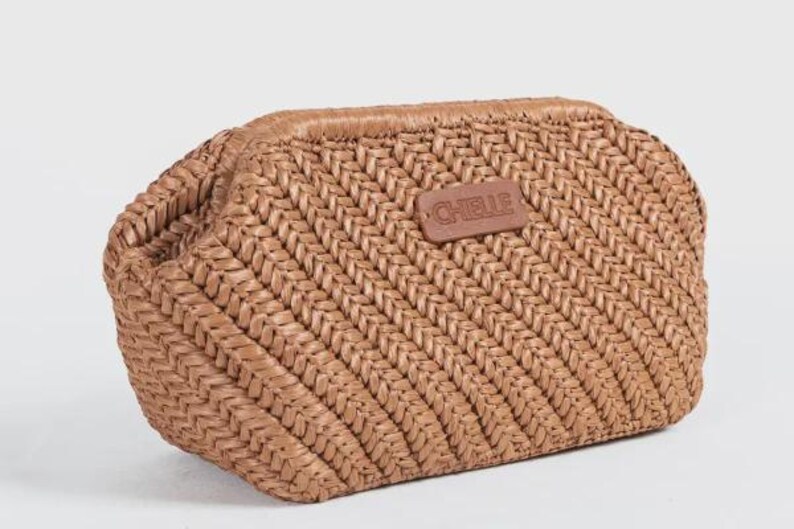 May include: A woven, light brown clutch bag with a rectangular shape and a rounded top. The bag features a textured, diagonal weave pattern and a small, brown leather tag with the word "CHELLE" embossed on it.