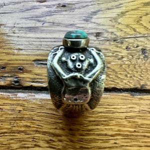May include: A small, silver-toned bottle with a turquoise stone on top. The bottle has a detailed, textured design featuring a stylized figure. The bottle is displayed on a wooden surface.