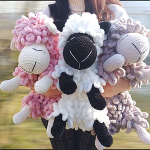 May include: Three crocheted stuffed sheep toys. One is pink, one is white with black face, and one is light gray.