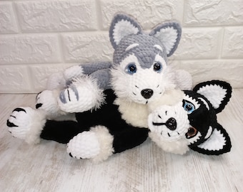 May include: Two crocheted husky dog plushies, one gray and white and one black and white, lying on a white wooden surface.