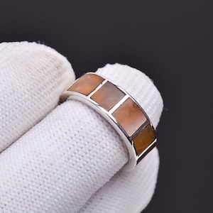 May include: A silver ring with a band featuring a series of square, light brown inlays. The ring is held by a gloved hand, showcasing the jewelry's design and craftsmanship. The inlays have a subtle, natural color variation.