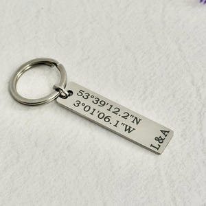 Könnte beinhalten: Ein rechteckiger Schlüsselanhänger in Silber mit einem geteilten Ring. Der Schlüsselanhänger ist mit Koordinaten und den Initialen "L&A" graviert. Der Hintergrund ist eine weiße, strukturierte Oberfläche.