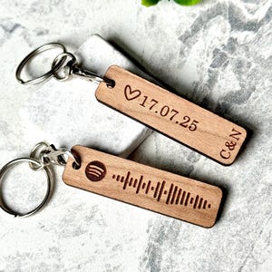 May include: Two wooden keyrings with silver rings. One keyring has a heart symbol, the date 17.07.25, and the initials C&N. The other keyring has a Spotify code.