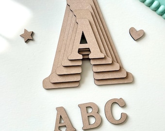 Puede incluir: Letra "A" de madera en un diseño en capas, con letras de madera adicionales "B" y "C", una estrella y un corazón. Las letras son de color marrón claro y están dispuestas sobre una superficie blanca. La imagen está bien iluminada y es adecuada para proyectos de manualidades.