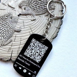 May include: Black metal keychain with a silver chain and a black tag featuring a QR code and the text "All I Want for Christmas Is You Mariah Carey". The tag also has music player icons.