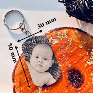 Puede incluir: Un llavero personalizado con una imagen en escala de grises de un beb&eacute;. El llavero mide 5 cm de largo y 3 cm de ancho, sujeto a un llavero plateado. El fondo es una superficie de madera con detalles de pan de oro.