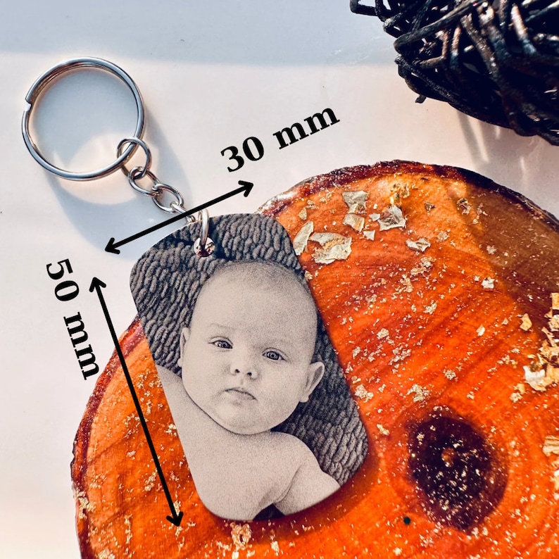 Puede incluir: Un llavero personalizado con una imagen en escala de grises de un beb&eacute;. El llavero mide 5 cm de largo y 3 cm de ancho, sujeto a un llavero plateado. El fondo es una superficie de madera con detalles de pan de oro.