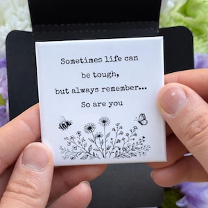 May include: A white square card with the text "Sometimes life can be tough, but always remember... So are you." The card features a bee and butterfly illustration with floral accents. The card is held between two hands.
