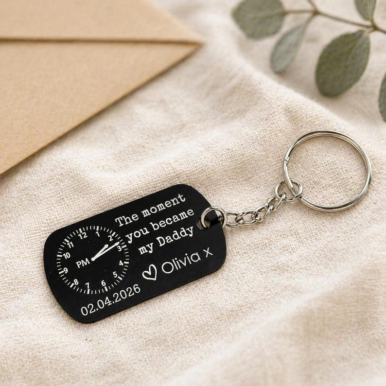 K&ouml;nnte beinhalten: Schwarzer rechteckiger Schl&uuml;sselanh&auml;nger mit einem Zifferblatt-Design und dem Text "The moment you became my Daddy" und "Olivia X". Das Datum "02.04.2026" ist ebenfalls angegeben. Er hat eine silberne Kette und einen Ring.