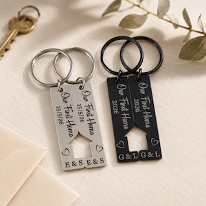 May include: Two keyrings with the text "Our First Home" and the year 2026. One keyring is silver with the date 15/5/26 and initials "E & S & S". The other is black with initials "G & L & L".
