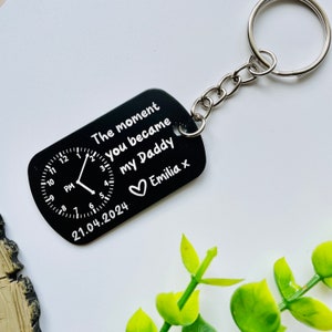 Puede incluir: Llavero rectangular negro con un anillo plateado. El llavero presenta texto blanco: "The moment you became my Daddy Emilia x" y un dise&ntilde;o de reloj. Tambi&eacute;n se muestra la fecha "21.04.2024".