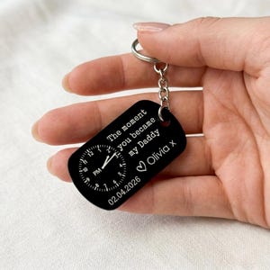 K&ouml;nnte beinhalten: Schwarzer rechteckiger Schl&uuml;sselanh&auml;nger mit einem Zifferblatt-Design und dem Text "The moment you became my Daddy Olivia x 02.04.2026". Der Schl&uuml;sselanh&auml;nger hat eine silberne Kette und einen Ring.