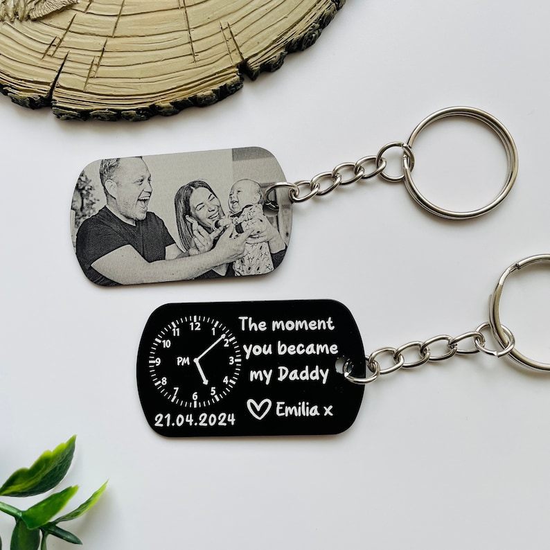 Puede incluir: Dos llaveros con cadenas y anillos plateados. Uno presenta una foto en blanco y negro de una familia. El otro es negro con un dise&ntilde;o de reloj y el texto "The moment you became my Daddy Emilia x" y la fecha 21.04.2024.