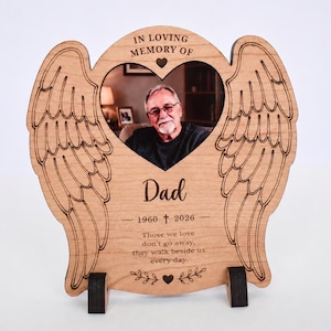 May include: Wooden memorial photo frame shaped like angel wings. The frame holds a photo of a man and includes the text "IN LOVING MEMORY OF Dad 1960 + 2026". The bottom of the frame has the text "Those we love don't go away, they walk beside us every day."