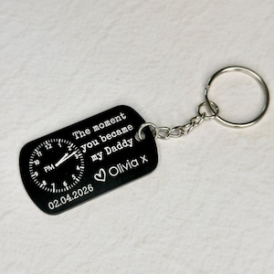 K&ouml;nnte beinhalten: Schwarzer rechteckiger Schl&uuml;sselanh&auml;nger mit Zifferblatt-Design und dem Text "The moment you became my Daddy Olivia x 02.04.2026". Der Schl&uuml;sselanh&auml;nger ist an einer silbernen Kette und einem Schl&uuml;sselring befestigt.