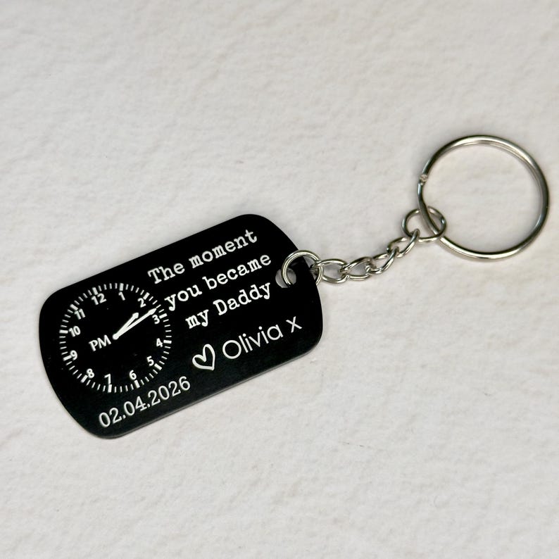 K&ouml;nnte beinhalten: Schwarzer rechteckiger Schl&uuml;sselanh&auml;nger mit Zifferblatt-Design und dem Text "The moment you became my Daddy Olivia x 02.04.2026". Der Schl&uuml;sselanh&auml;nger ist an einer silbernen Kette und einem Schl&uuml;sselring befestigt.