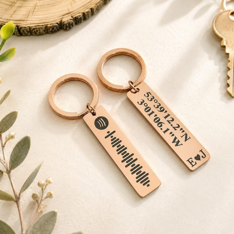 Op de afbeelding: Twee ros&eacute;gouden sleutelhangers met rechthoekige labels. E&eacute;n label heeft een Spotify-code, de andere heeft co&ouml;rdinaten en initialen. De sleutelhangers liggen op een licht oppervlak met een sleutel en groen in de achtergrond.
