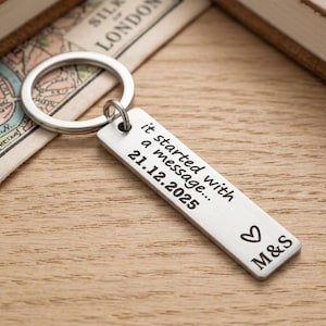 Op de afbeelding: Een zilveren rechthoekige sleutelhanger met de tekst "it started with a message... 21.12.2025" en initialen "M&S" met een hartsymbool. De sleutelhanger ligt op een houten oppervlak met een kaart en een boek op de achtergrond.
