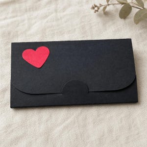 K&ouml;nnte beinhalten: Ein schwarzer Papierumschlag mit einem roten herzf&ouml;rmigen Akzent auf der Vorderseite. Der Umschlag hat einen gebogenen Klappenverschluss. Der Hintergrund ist ein neutraler, cremefarbener Stoff.