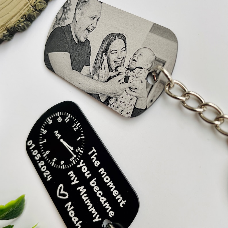 Puede incluir: Dos llaveros. Un llavero rectangular con una foto en blanco y negro de una familia. El otro es negro con un dise&ntilde;o de reloj y el texto "The moment you became my Mummy Noah".