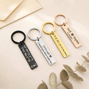 Op de afbeelding: Vier sleutelhangers met rechthoekige hangers. E&eacute;n zwarte sleutelhanger heeft co&ouml;rdinaten en initialen. Een zilveren sleutelhanger heeft de tekst "Drive safe I need you here with me". Een gouden sleutelhanger heeft initialen en een datum.