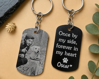 May include: Two keychains with silver rings. One keychain has a black and white photo of a man and a dog. The other keychain is black with the text "Once by my side, forever in my heart" and the name "Oscar".