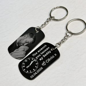 K&ouml;nnte beinhalten: Zwei schwarze Schl&uuml;sselanh&auml;nger mit silbernen Ketten und Ringen. Einer zeigt ein Graustufenbild eines Mannes und eines Babys. Der andere hat ein Zifferblatt-Design, den Text "The moment you became my Daddy Olivia x" und das Datum 02.04.2026.