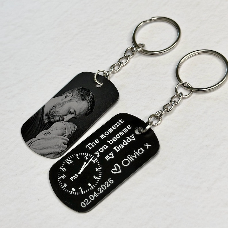 K&ouml;nnte beinhalten: Zwei schwarze Schl&uuml;sselanh&auml;nger mit silbernen Ketten und Ringen. Einer zeigt ein Graustufenbild eines Mannes und eines Babys. Der andere hat ein Zifferblatt-Design, den Text "The moment you became my Daddy Olivia x" und das Datum 02.04.2026.