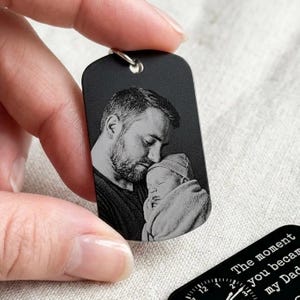 K&ouml;nnte beinhalten: Schwarzer Anh&auml;nger in Form einer Erkennungsmarke mit einer silbernen Schlaufe. Der Anh&auml;nger zeigt ein Schwarz-Wei&szlig;-Bild eines Mannes, der ein Baby h&auml;lt. Der Text "The moment you became my Dad" ist auf einem zweiten Anh&auml;nger zu sehen.