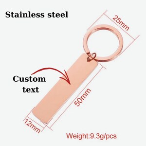 Op de afbeelding: Een ros&eacute;gouden roestvrijstalen sleutelhanger met een rechthoekige plaat voor aangepaste tekst. De plaat is 50 mm lang, 12 mm breed en de ring heeft een diameter van 25 mm. De sleutelhanger weegt 9,3 g. De tekst "Stainless steel" en "Custom text" zijn ook zichtbaar.