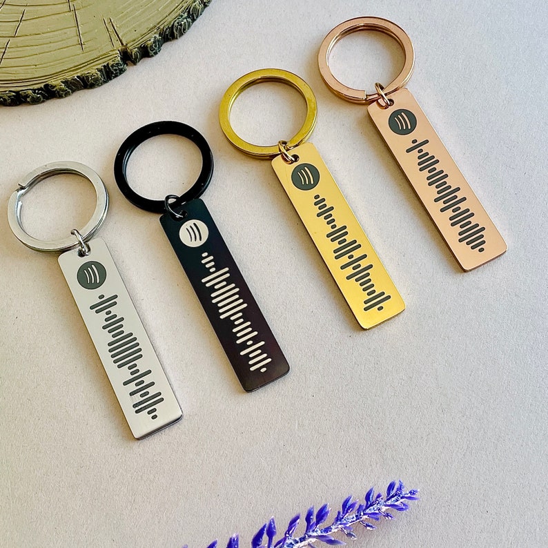 Four rectangular personalised metal Spotify code keyrings in silver, black, gold and rose gold, laid on a light surface, each engraved with a name and scannable Spotify code, with a small purple sprig and wood slice in frame.