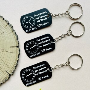 Puede incluir: Llaveros rectangulares negros con llaveros plateados. Cada llavero presenta un dise&ntilde;o de reloj y el texto "The moment you became my Daddy/Mummy/Grandma" con un nombre y una fecha. Los llaveros est&aacute;n conectados a una cadena plateada.