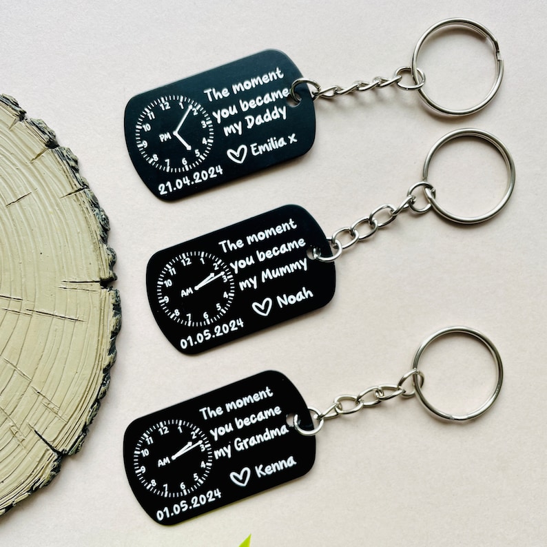 Puede incluir: Llaveros rectangulares negros con llaveros plateados. Cada llavero presenta un dise&ntilde;o de reloj y el texto "The moment you became my Daddy/Mummy/Grandma" con un nombre y una fecha. Los llaveros est&aacute;n conectados a una cadena plateada.