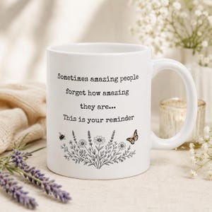 Op de afbeelding: Een witte keramische mok met zwarte tekst die luidt: Sometimes amazing people forget how amazing they are... This is your reminder. Onder de tekst staat een zwarte lijntekening van wilde bloemen, een kleine bij en een vlinder.