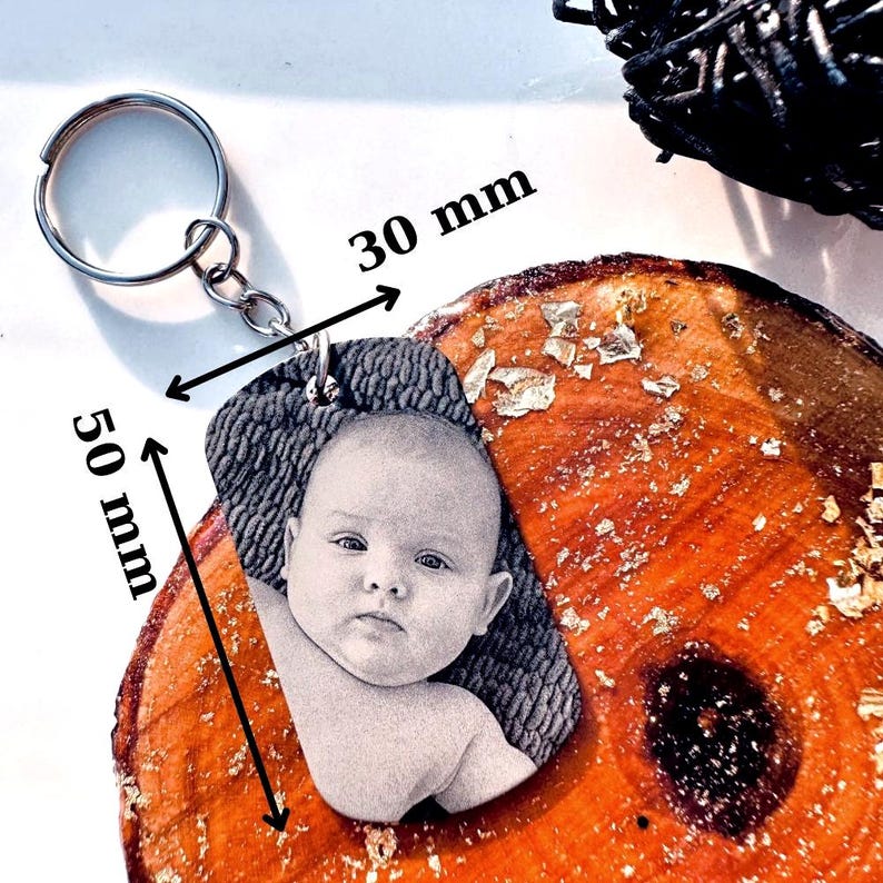 K&ouml;nnte beinhalten: Ein personalisierter Schl&uuml;sselanh&auml;nger mit einem Graustufenbild eines Babys. Der Anh&auml;nger ist 50 mm lang und 30 mm breit und an einem silbernen Schl&uuml;sselring befestigt. Der Hintergrund ist eine Holzoberfl&auml;che mit Goldblattakzenten.